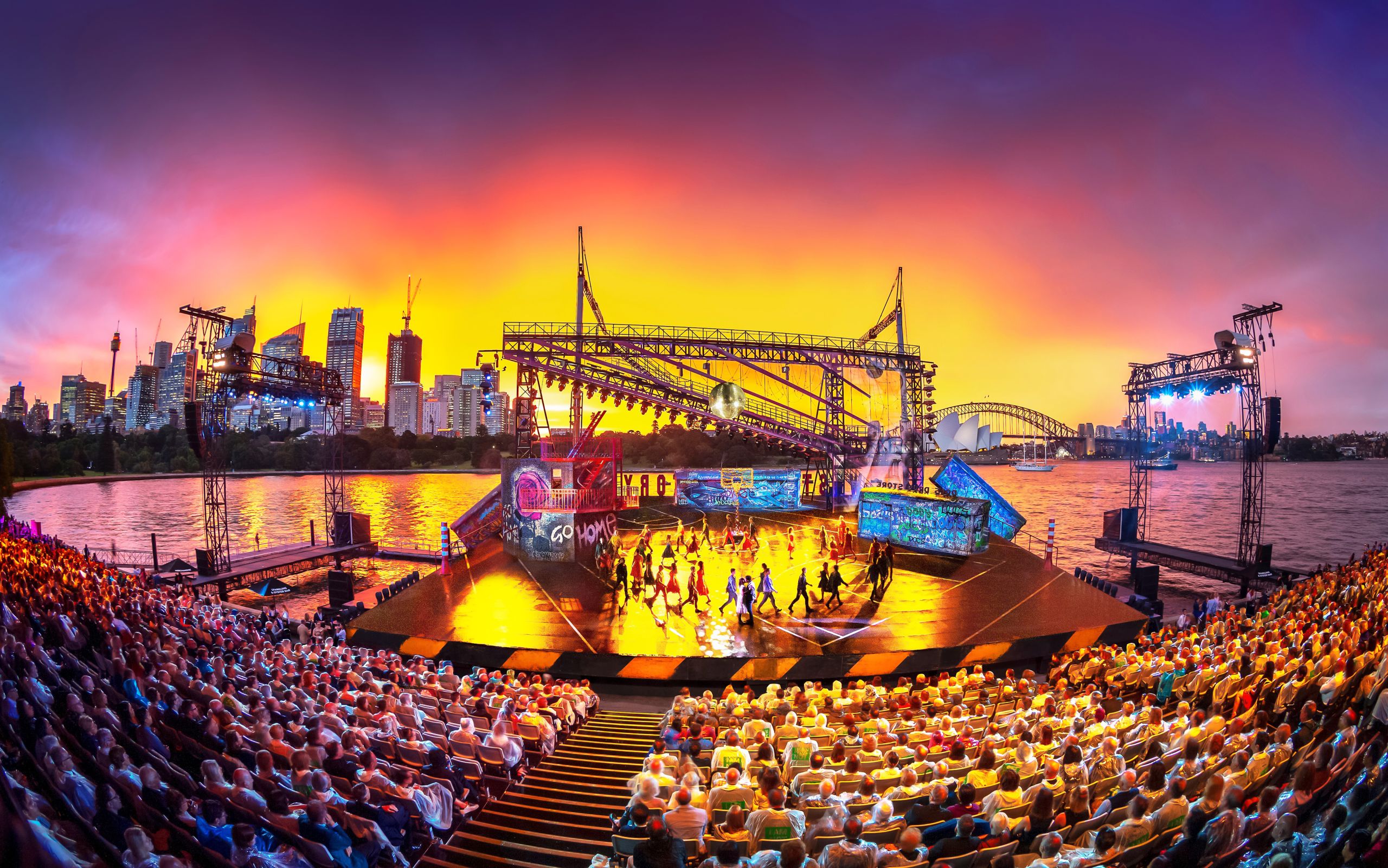 A large outdoor stage on Sydney Harbour, with a basketball court painted onto the surface, a freeway overpass and a mirrorball overhead.