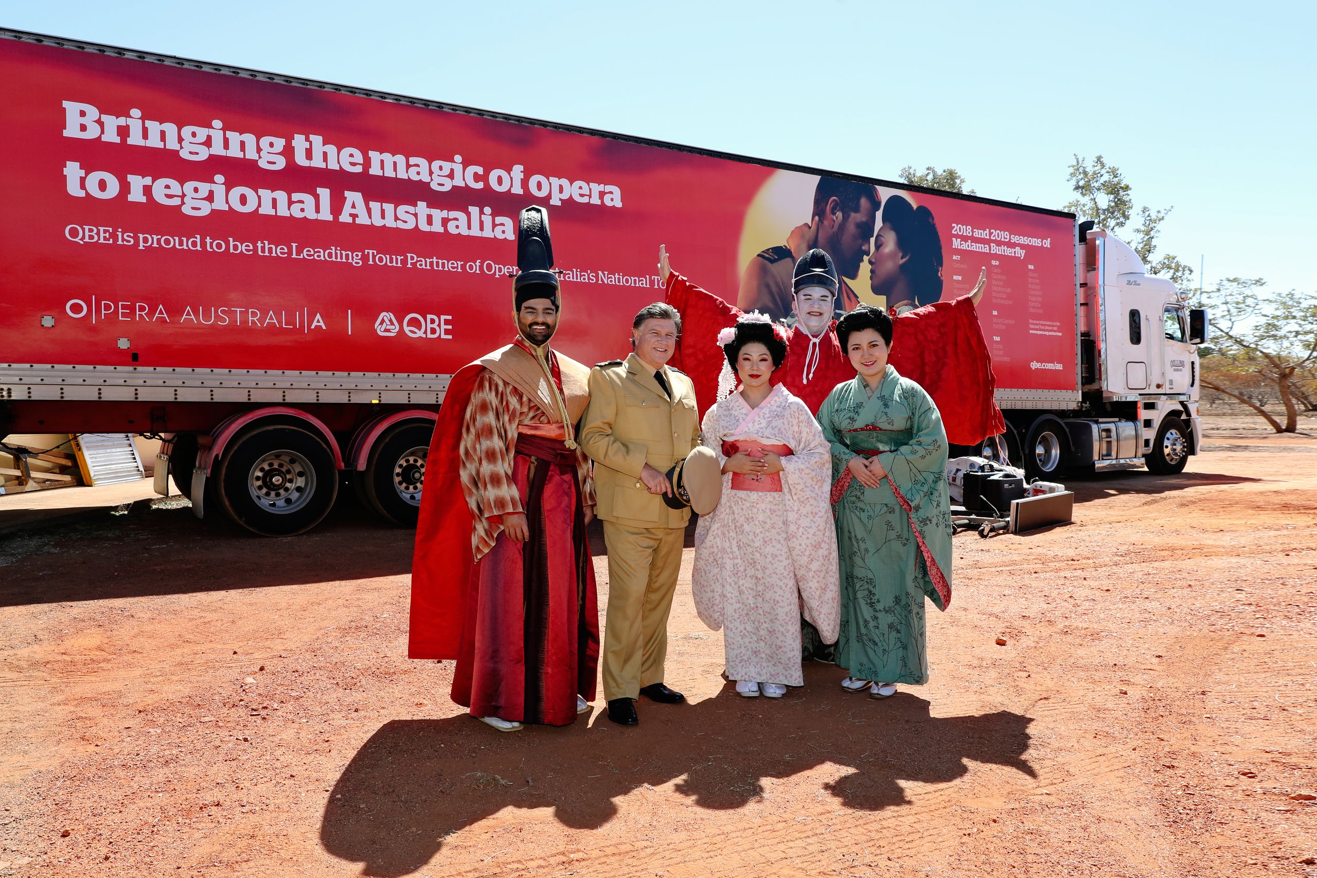 A group of singers in Japanese costumes stand in front of a large red truck which reads 'Bringing the magic of opera to regional Australia - QBE is proud to be the Leading Tour Partner of Opera Australia's National Tour'