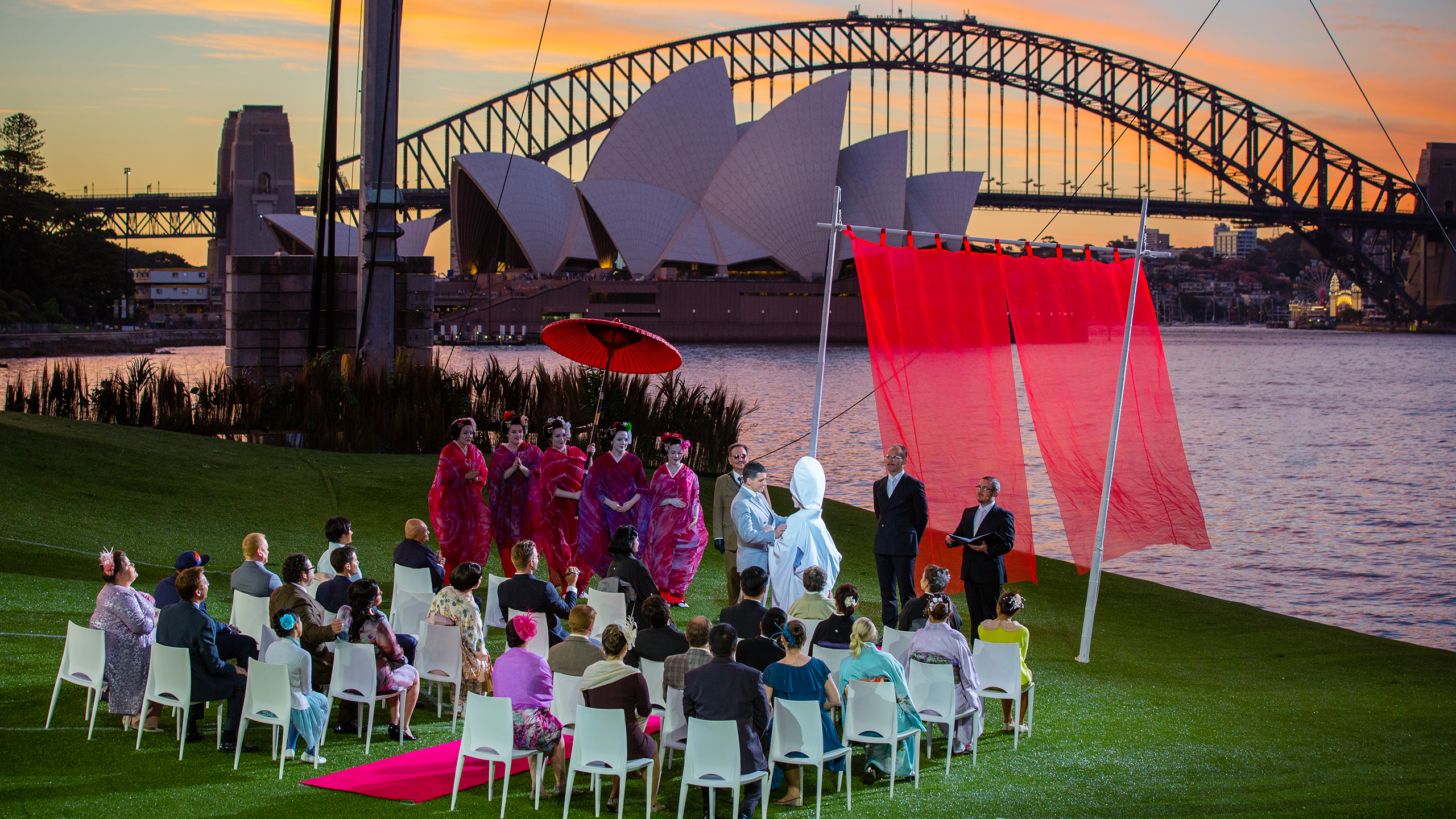 A wedding scene in a performance of Madama Butterfly on Sydney Harbour. The Sydney Harbour Bridge and Opera House appear behind a grassy hill and wedding guests.