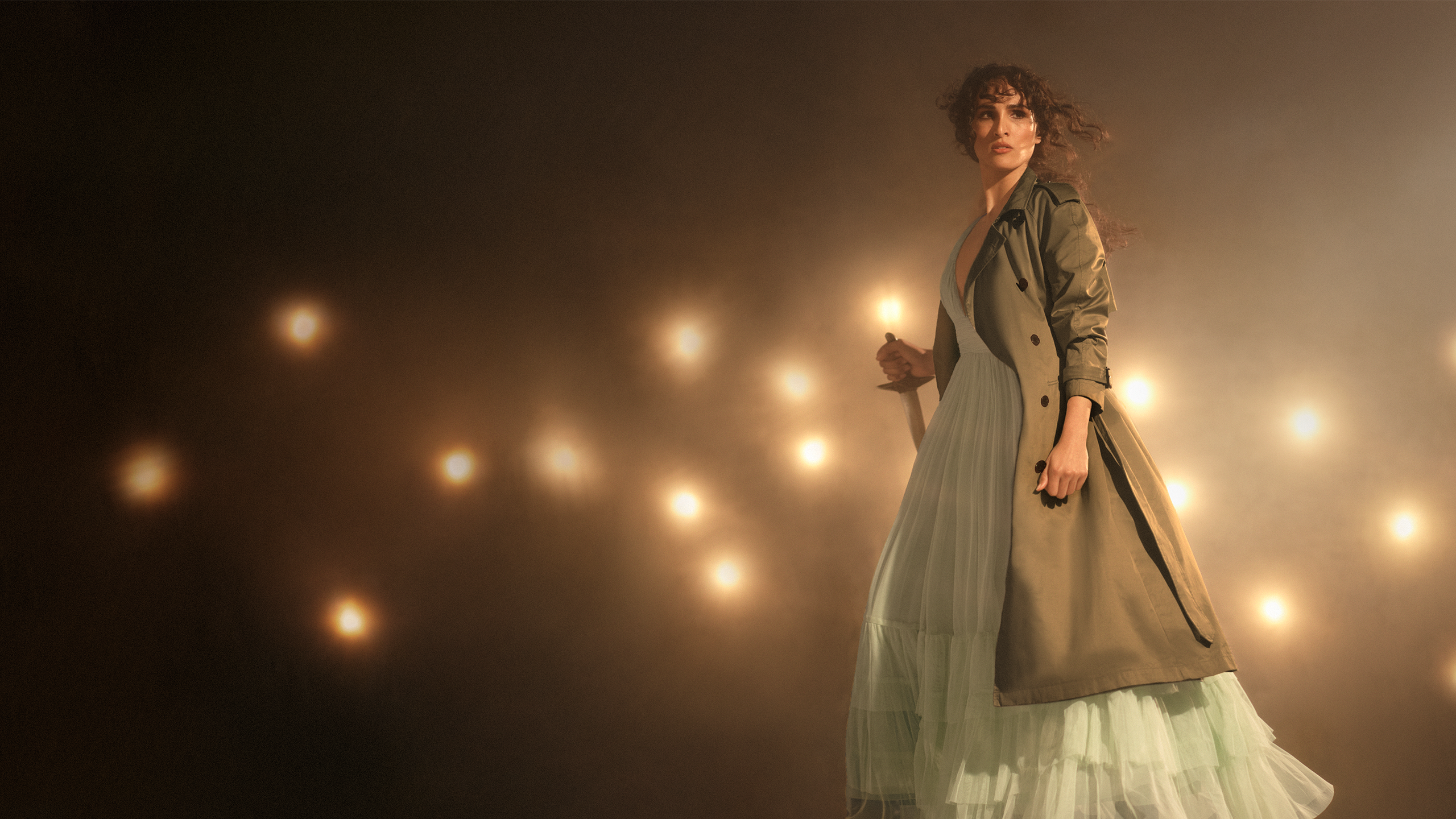 A woman stands in a trench coat, in front of bright spotlights, holding a dagger.