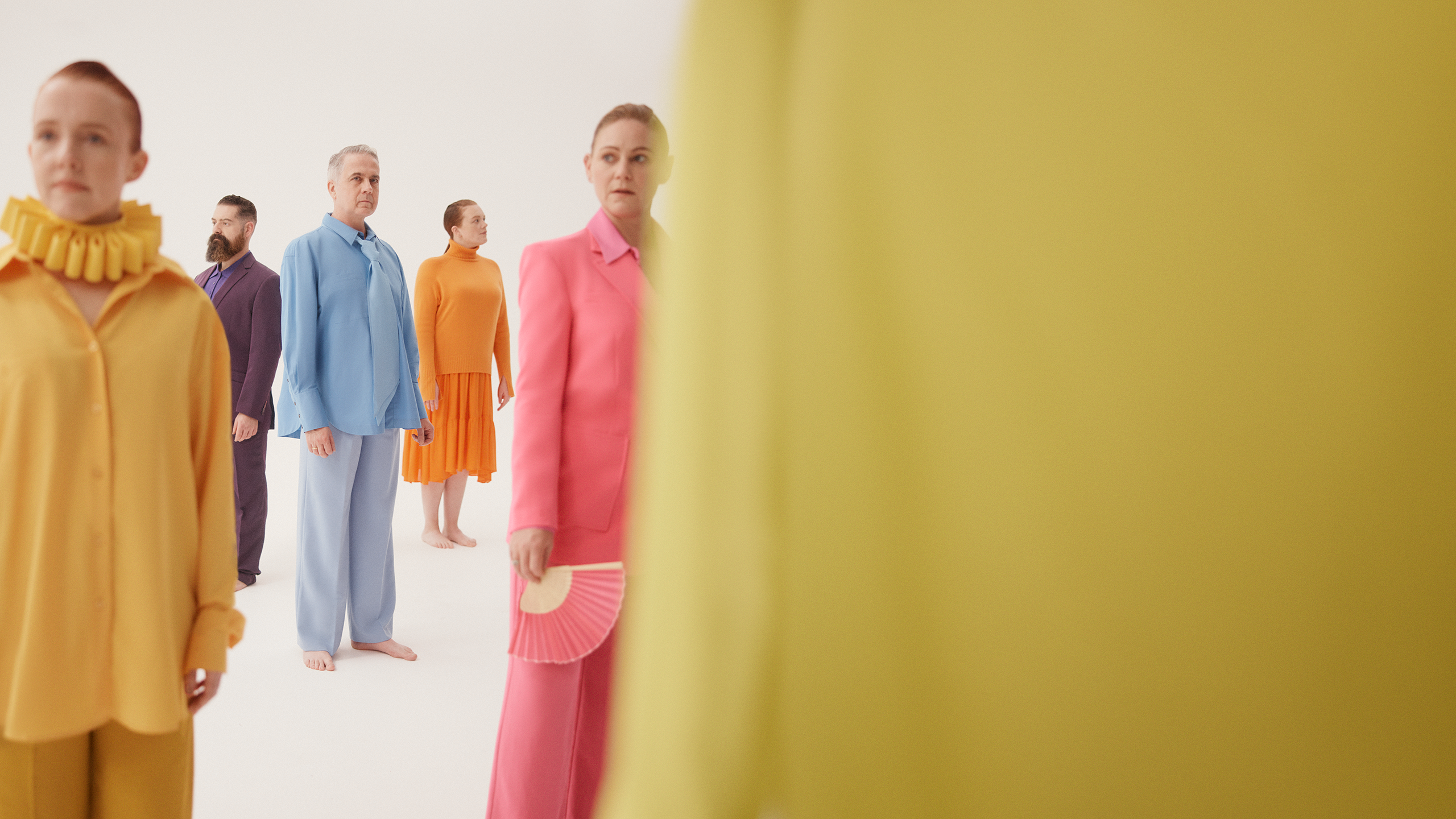 A group of Opera Australia Chorus members stand in brightly coloured costumes  in front of a white background.