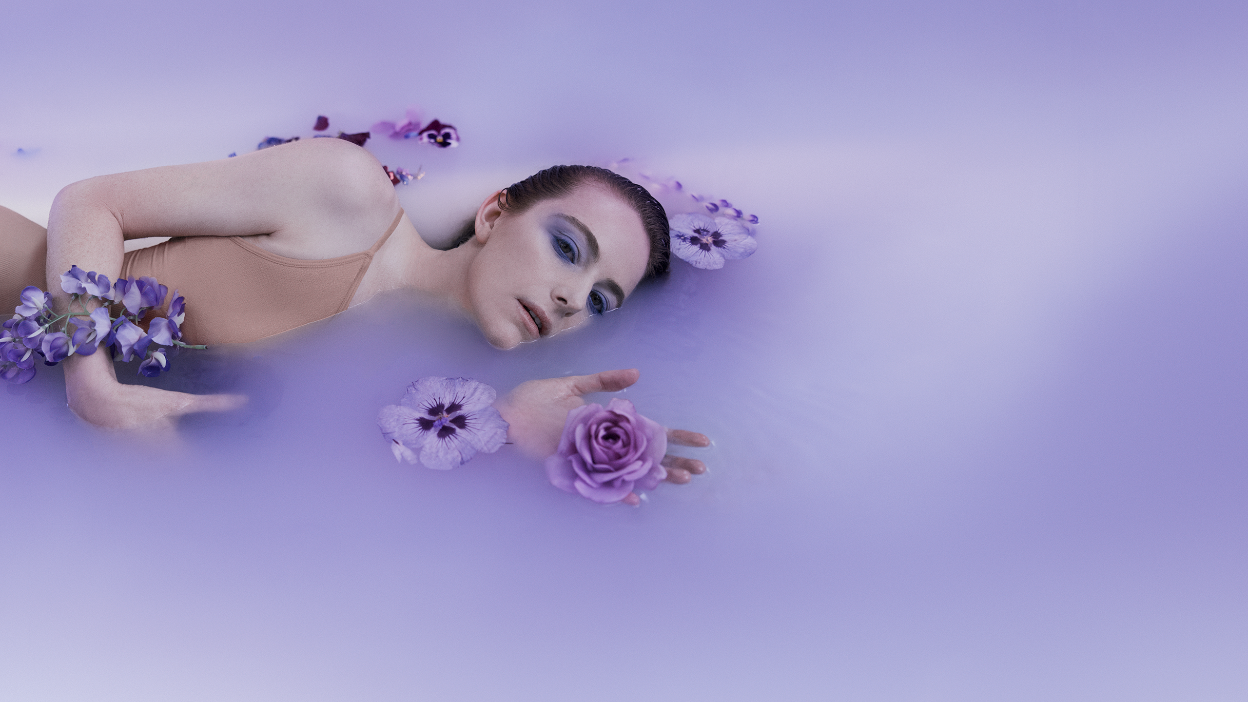 A woman lies in a milky pool of water, surrounded by purple flowers.