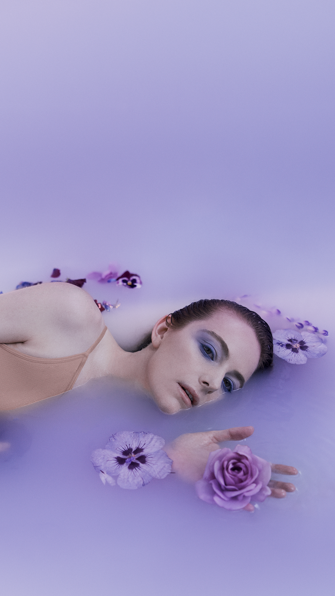 A woman lies in a milky pool of water, surrounded by purple flowers.