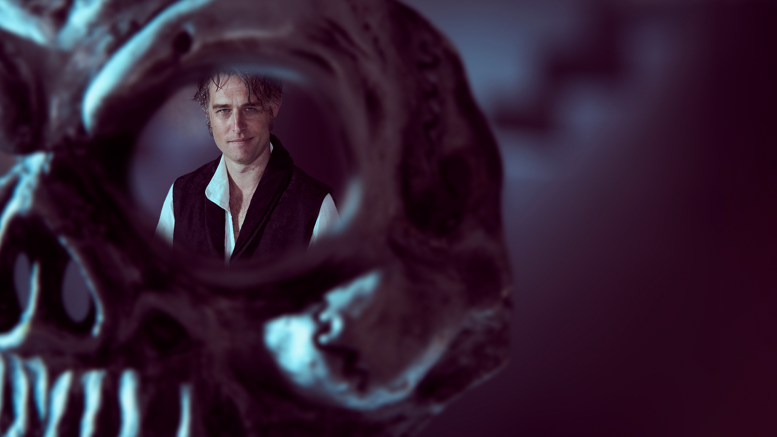 A man stares out from behind the eye of a dark grey skull.