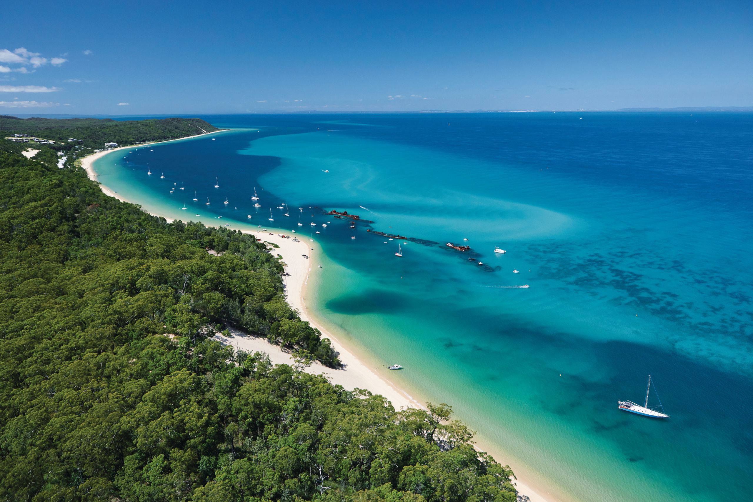 An aerial view of a coastline, with clear blue waters, white sand and green forests.