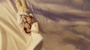 A man and a woman lie on a large canvas, painted as a dusky sky. They're gently embracing.