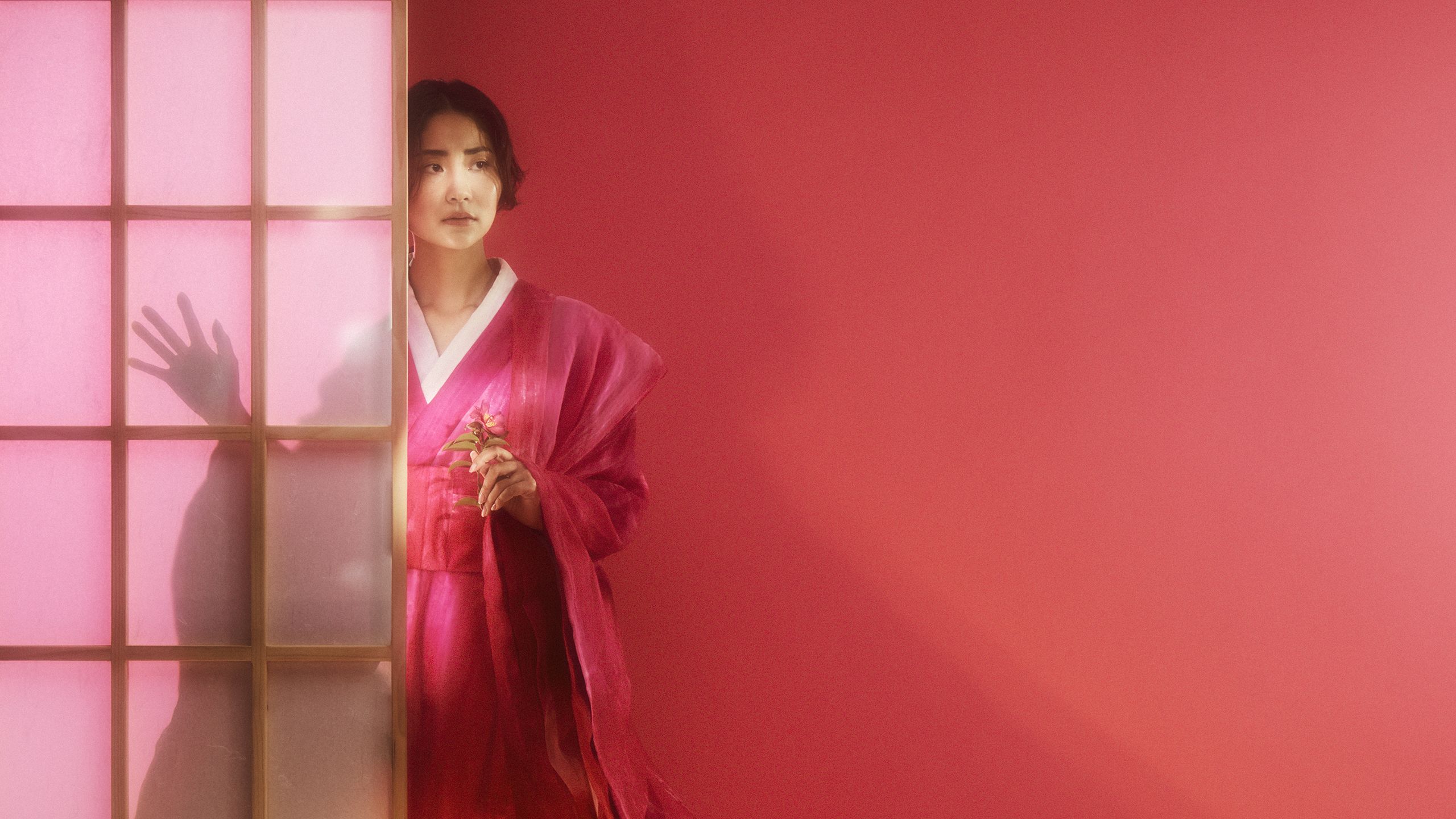 A portrait of a woman in a pink kimono. She's holding a flower and peeking around the corner of a shoji screen.