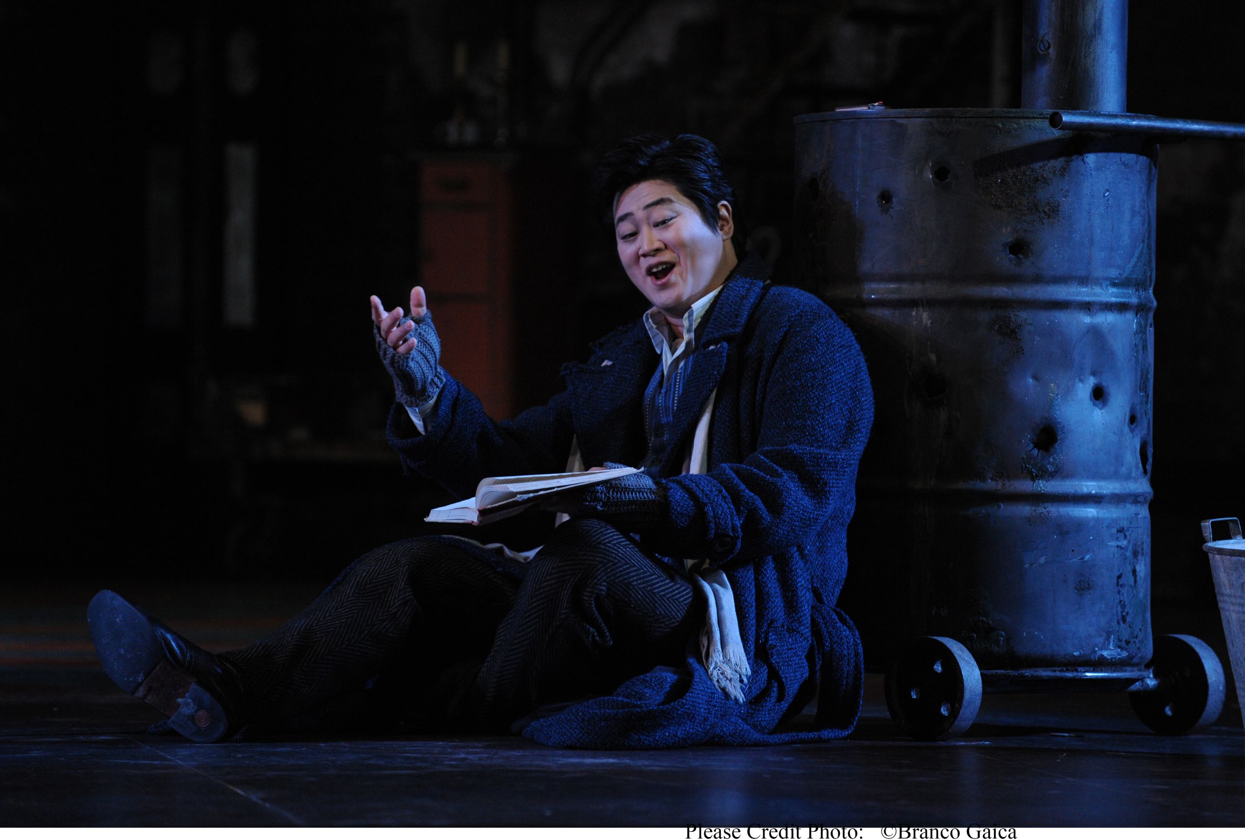 A man sits reading a book while dressed in warm winter clothes during a performance of La Boheme.