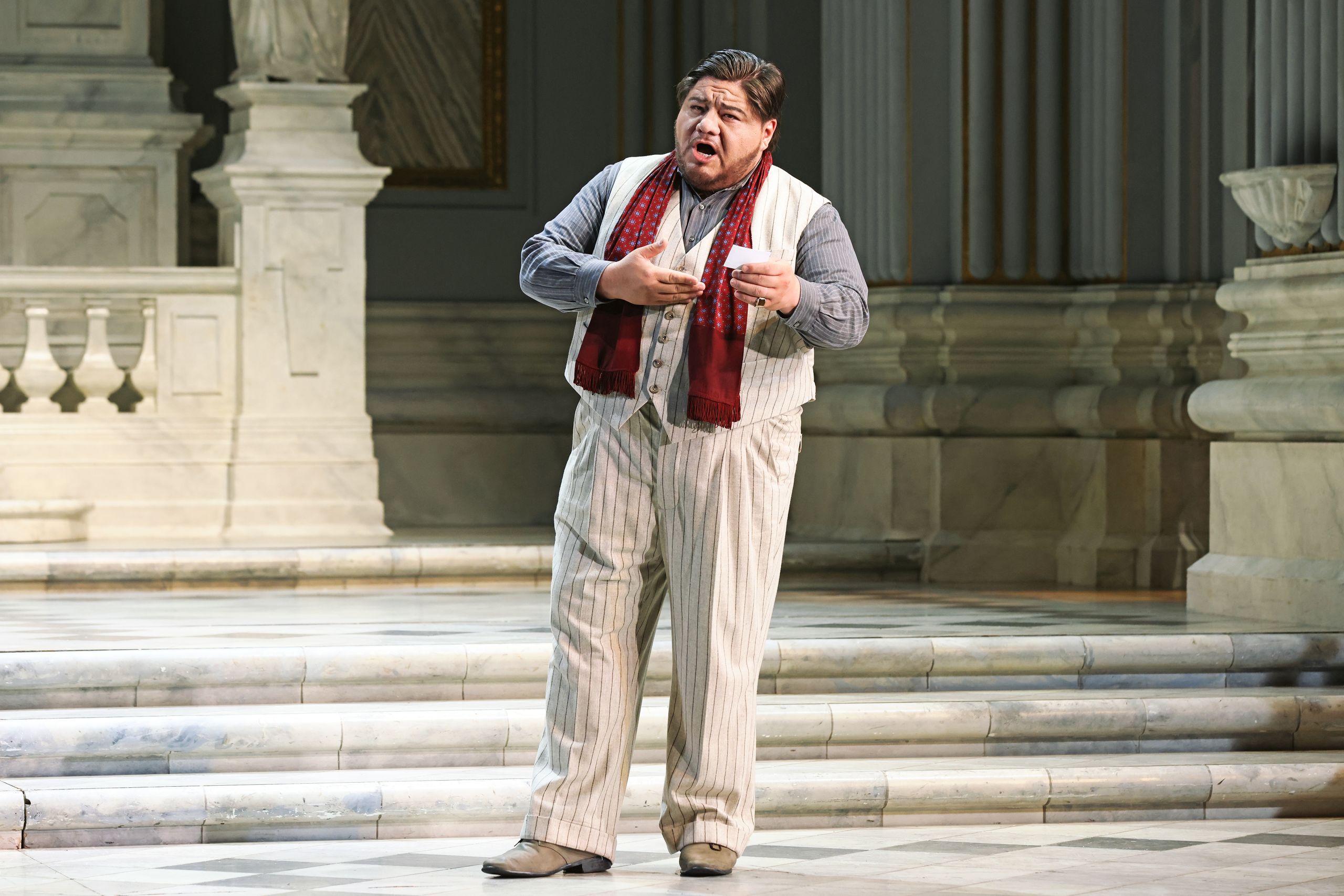 A man stands on a marble floor in a cream suit with a red scraf, reading a letter. He is in a performance of the opera Tosca.