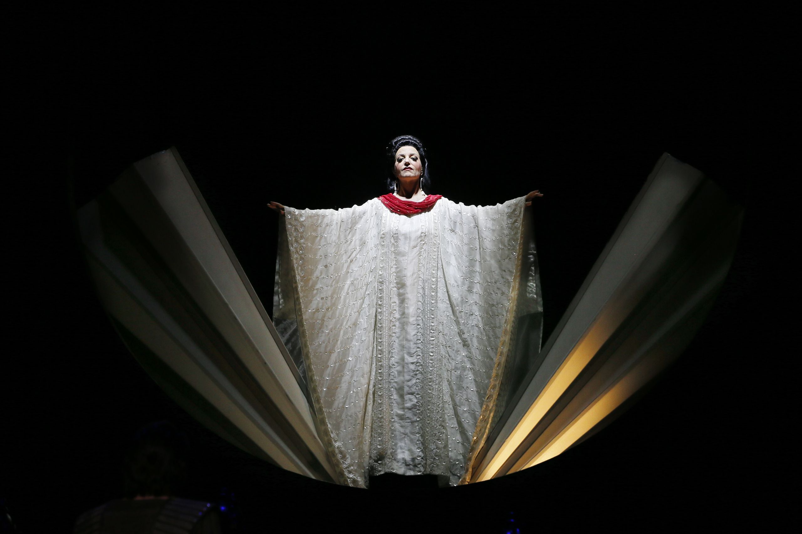 A woman wearing a long, flowing white kaftan stands with her arms held out wide in a performance of Turandot.