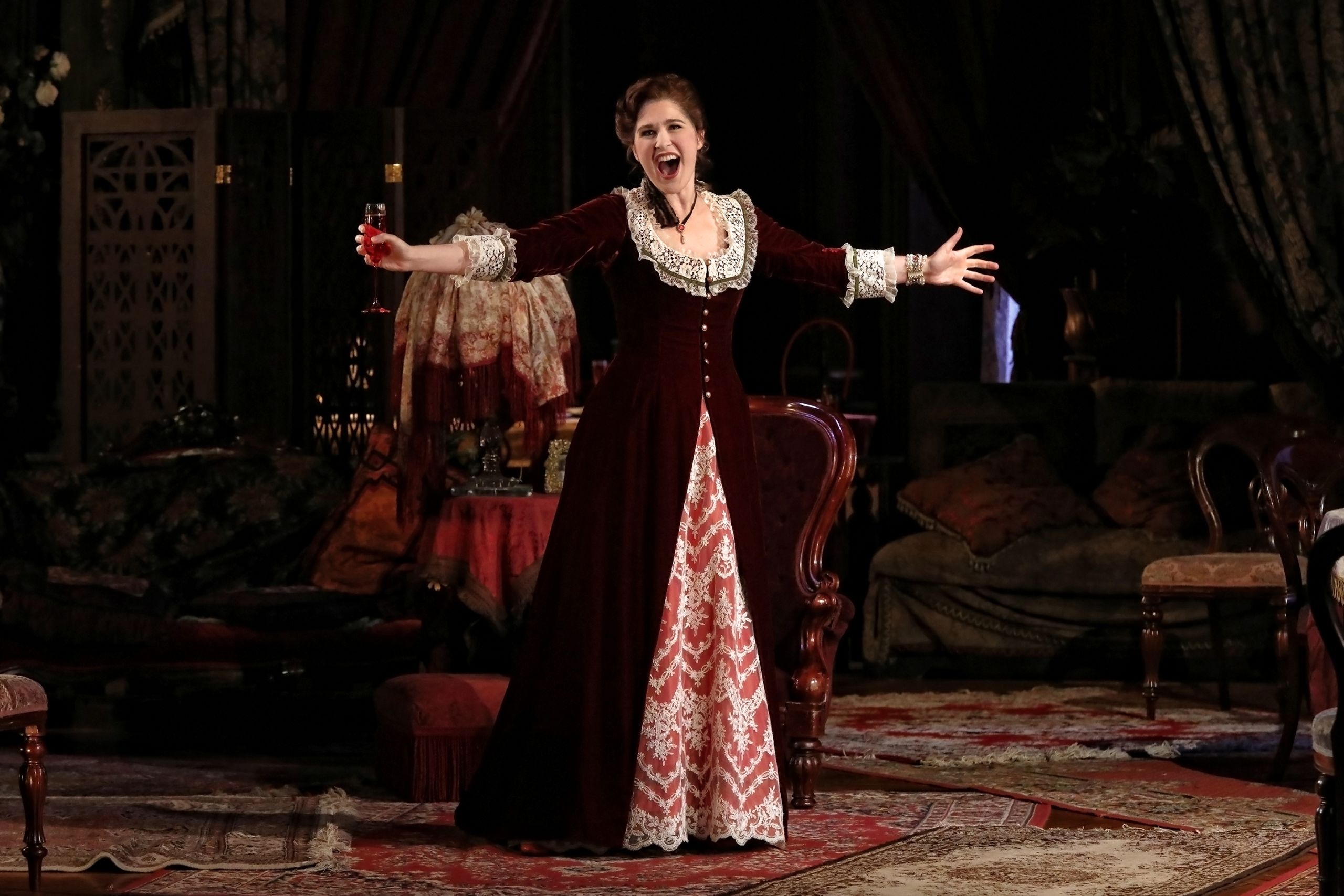 A woman stands in a red velvet dress holding a glass of champagne with her arms spread wide in a performance of La Traviata.