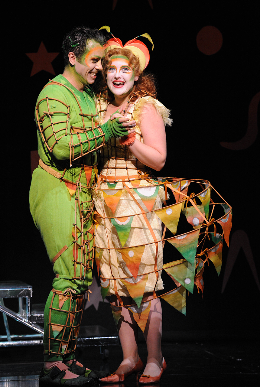 A man in a green costume with wicker details and orange makeup warmly embraces a woman in similar makeup and a bird-like costume with a large hoop skirt.