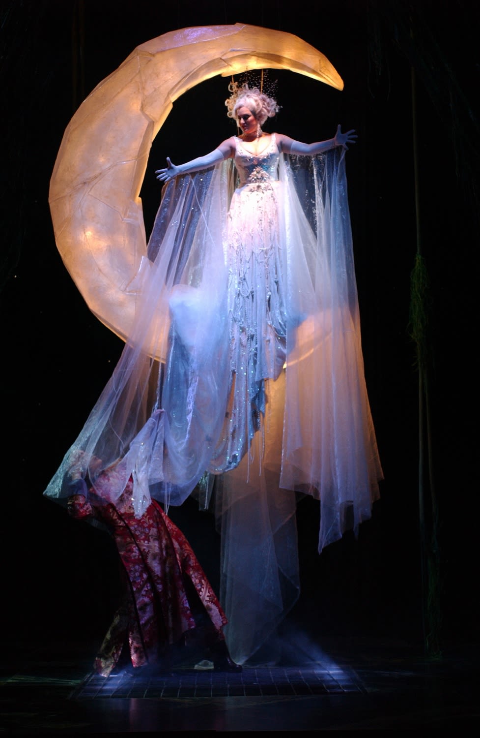 A woman in a long white gown is suspended from a white crescent moon above the stage of the Sydney Opera House.