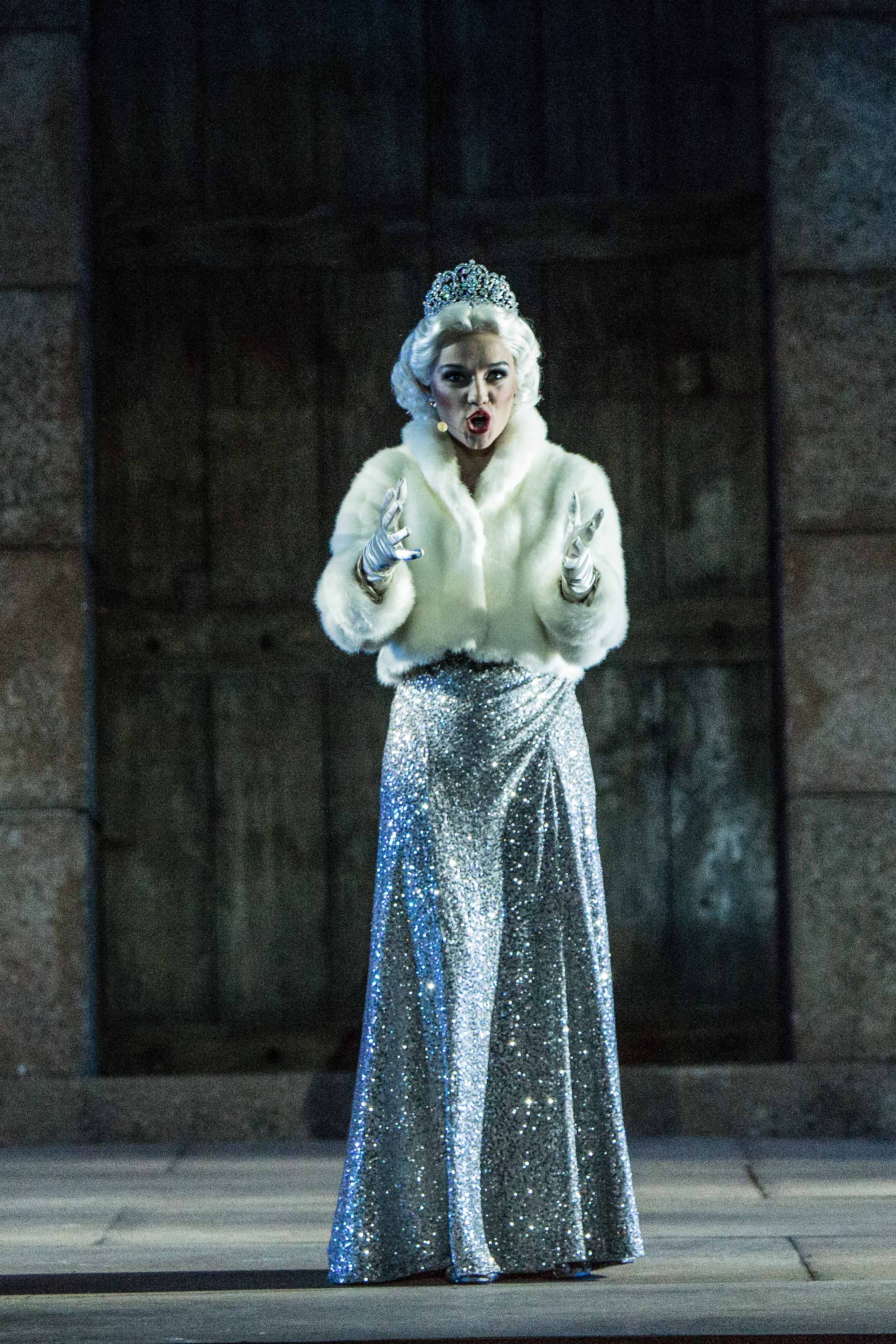 A female opera singer a silver sparkly gown with a white fur coat and a Marilyn Monroe-style wig stands holding her hands forward during a performance.