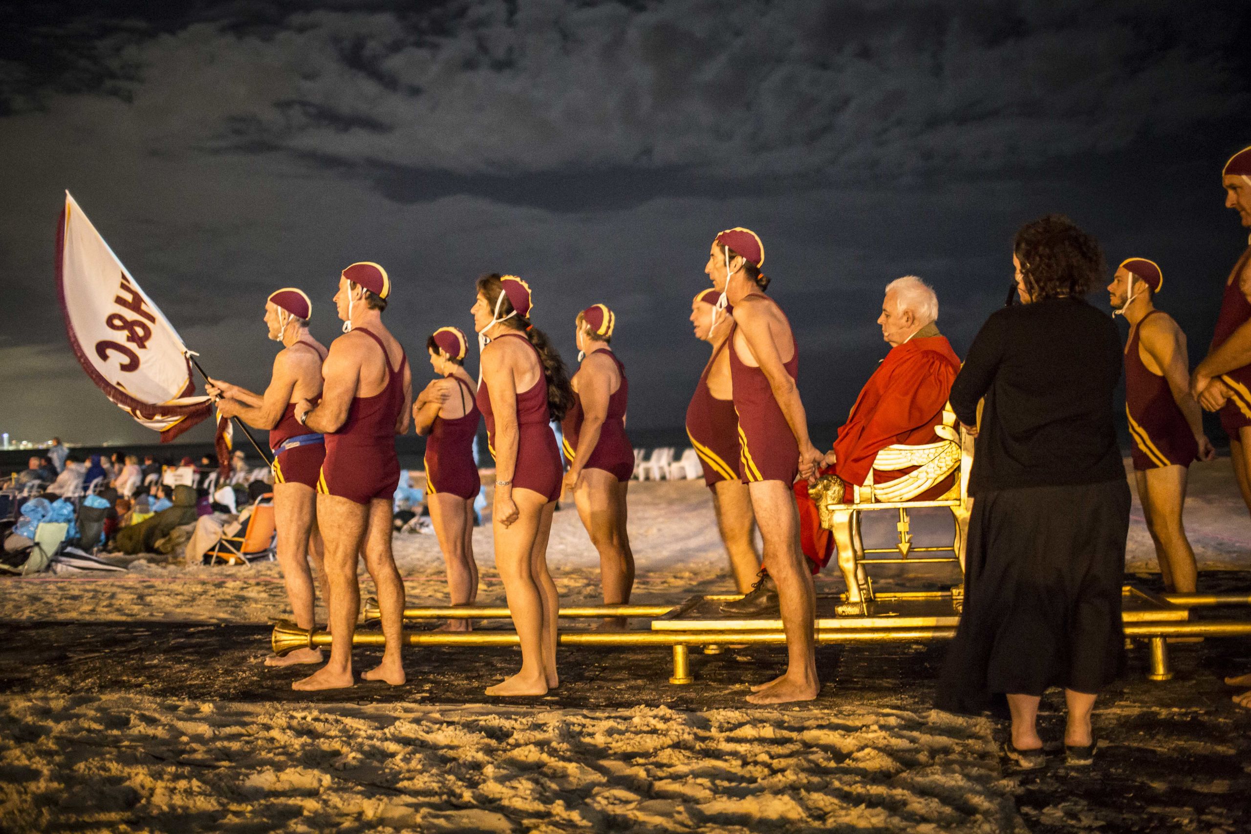 A group of surf lifesavers, in maroon one-piece swimsuits carry an older man in a large robe across the sand of a Gold Coast beach as part of a performance of The Magic Flute.