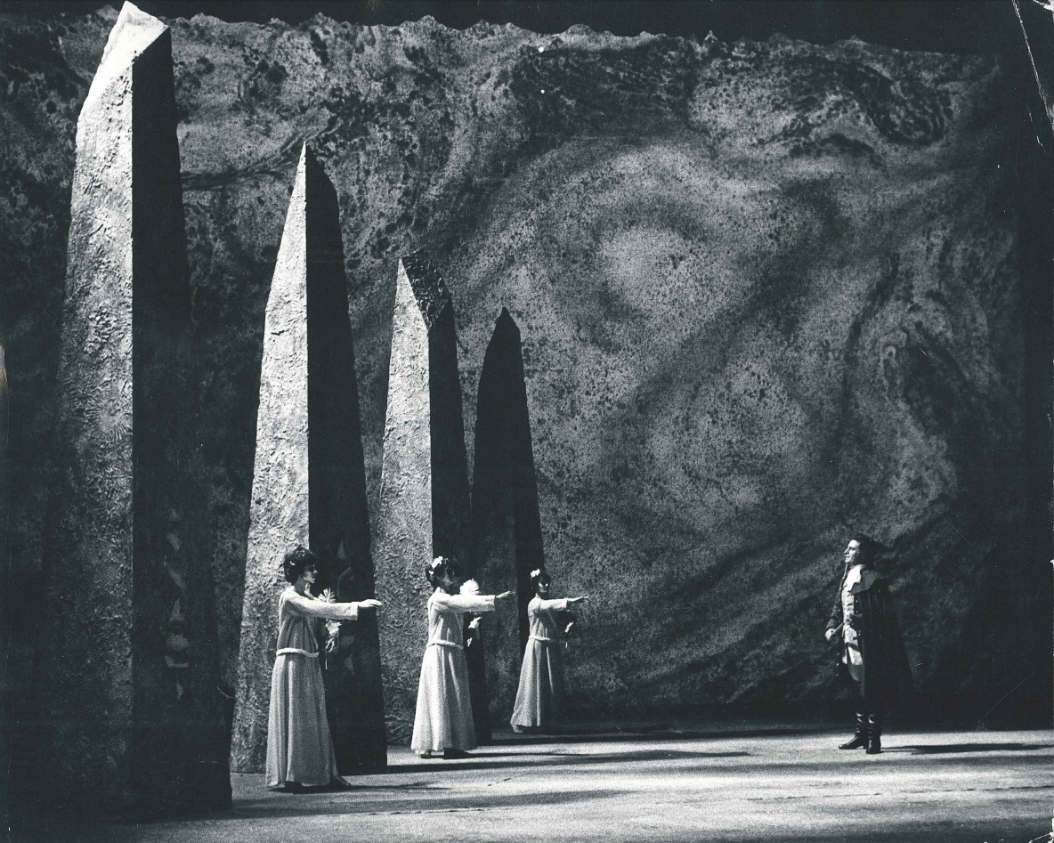 Three women in long robes stand in front of tall columns, holding their hands up at opera singer Anson Austin, in a performance of The Magic Flute.