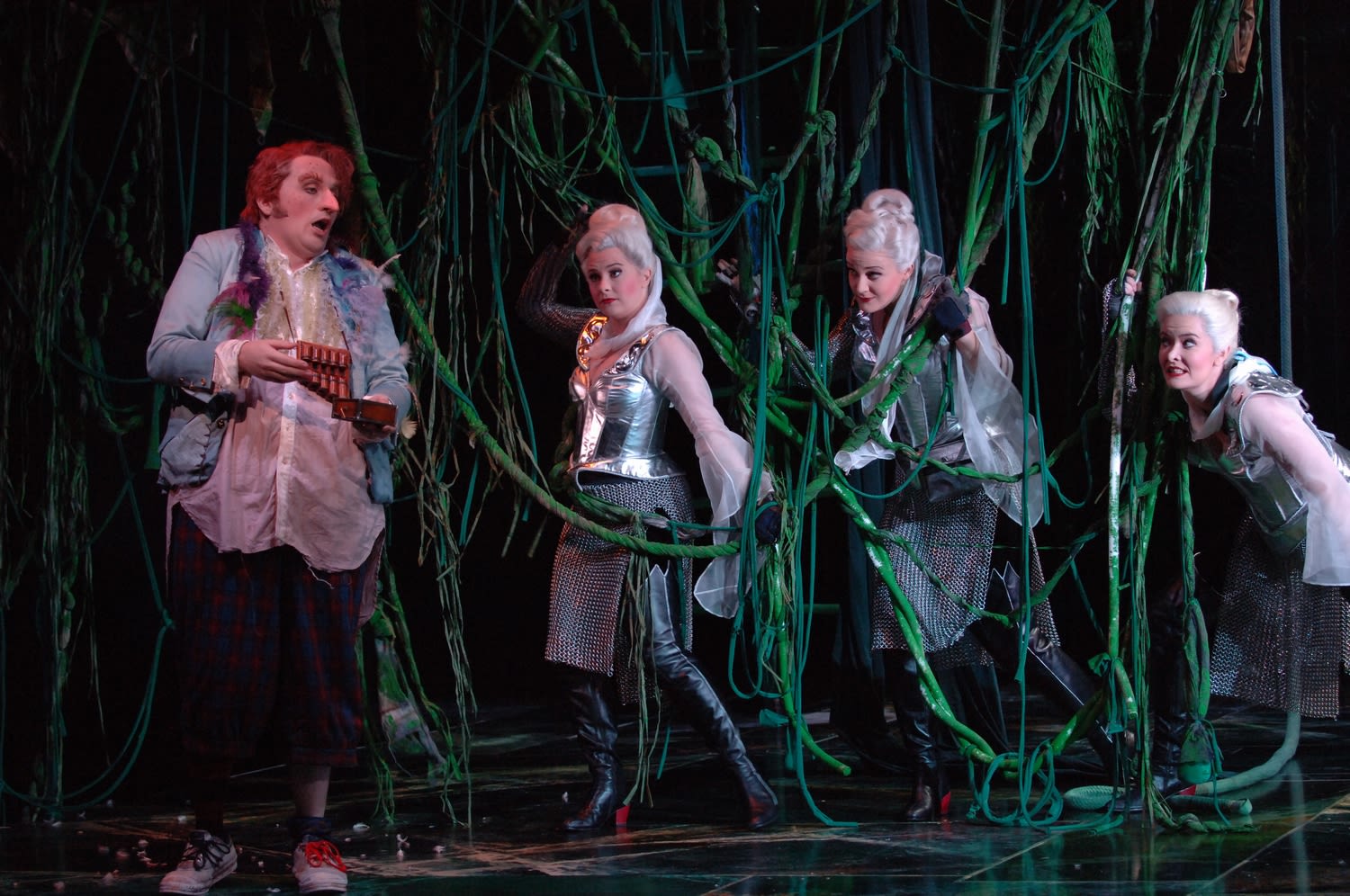 Opera singer Warwick Fyfe stands holding a panpipe, wearing a large fake nose. He is standing next to three women with white hair, who are surrounded by green vines.