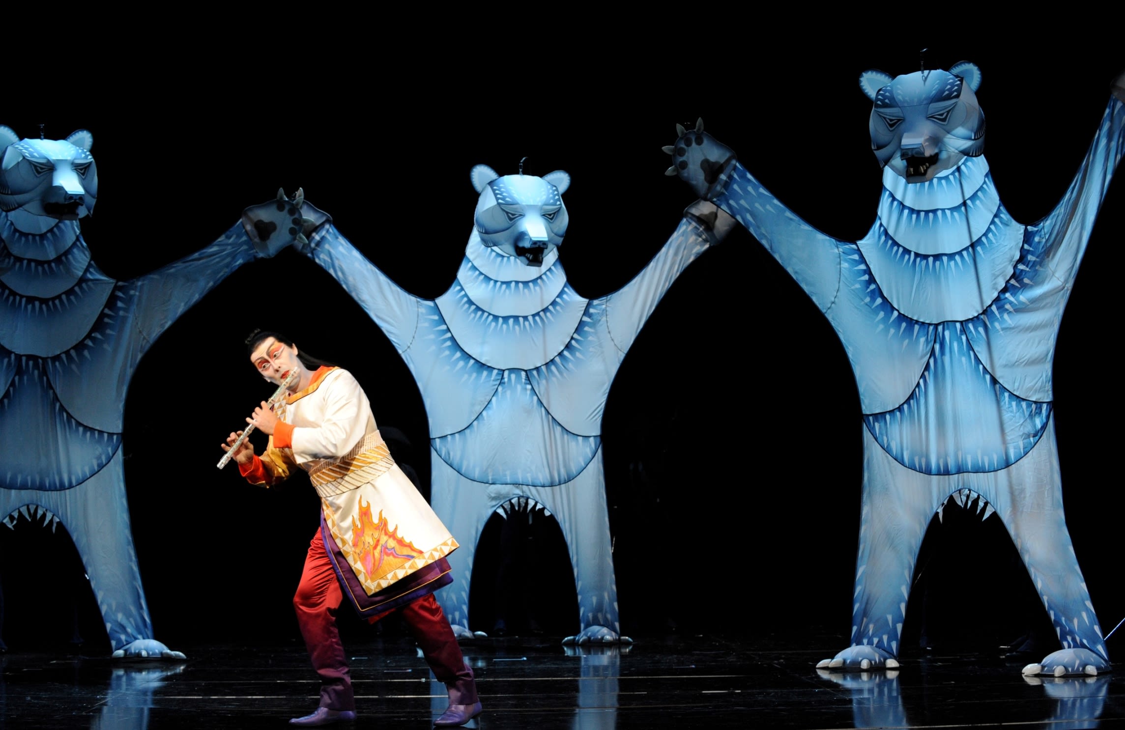 A man plays a flute while three large polar bear puppets tower over him in a performance of The Magic Flute.