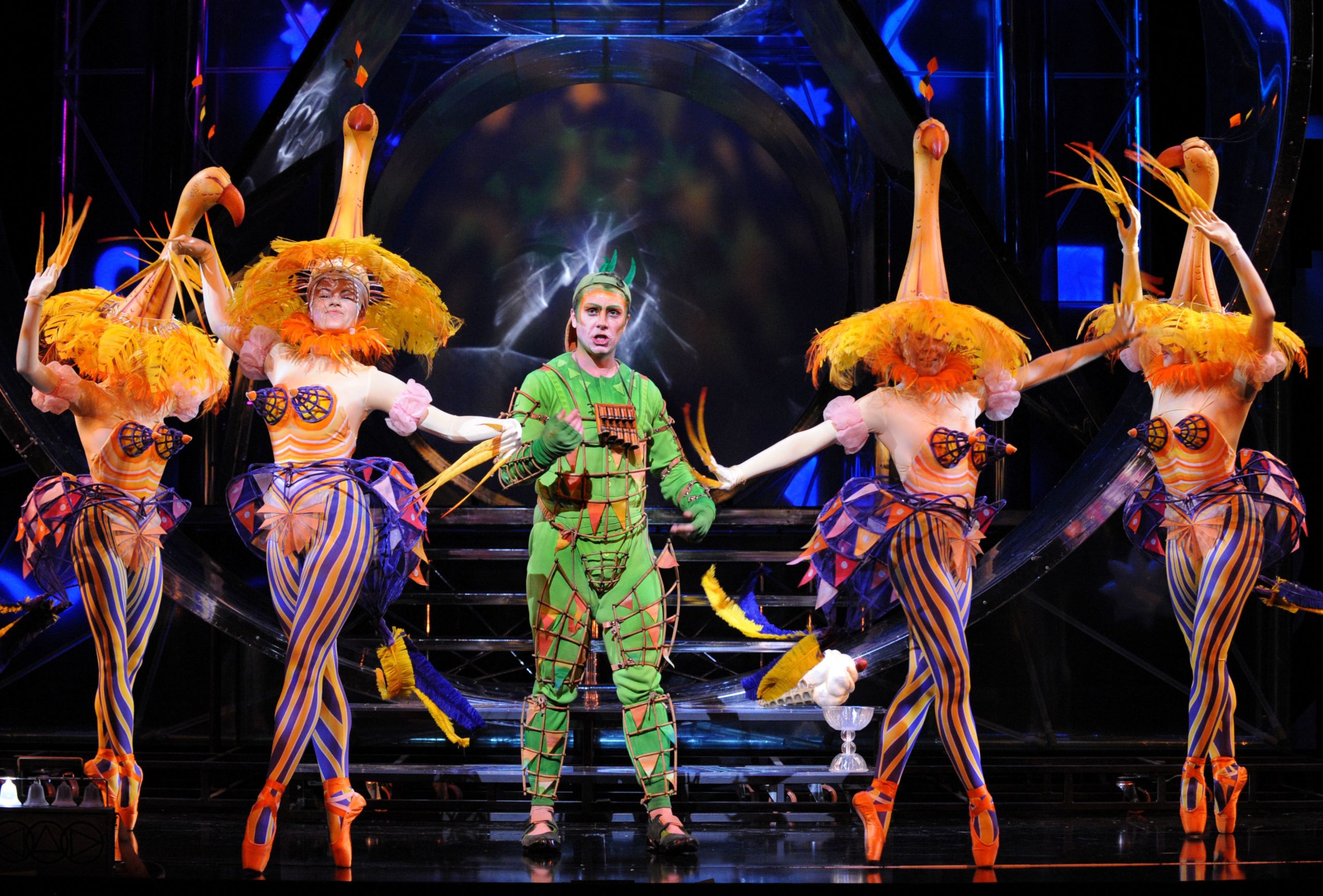 A man in a green costume is surrounded by four dancers in pointe shoes, who are costumed as peacocks.