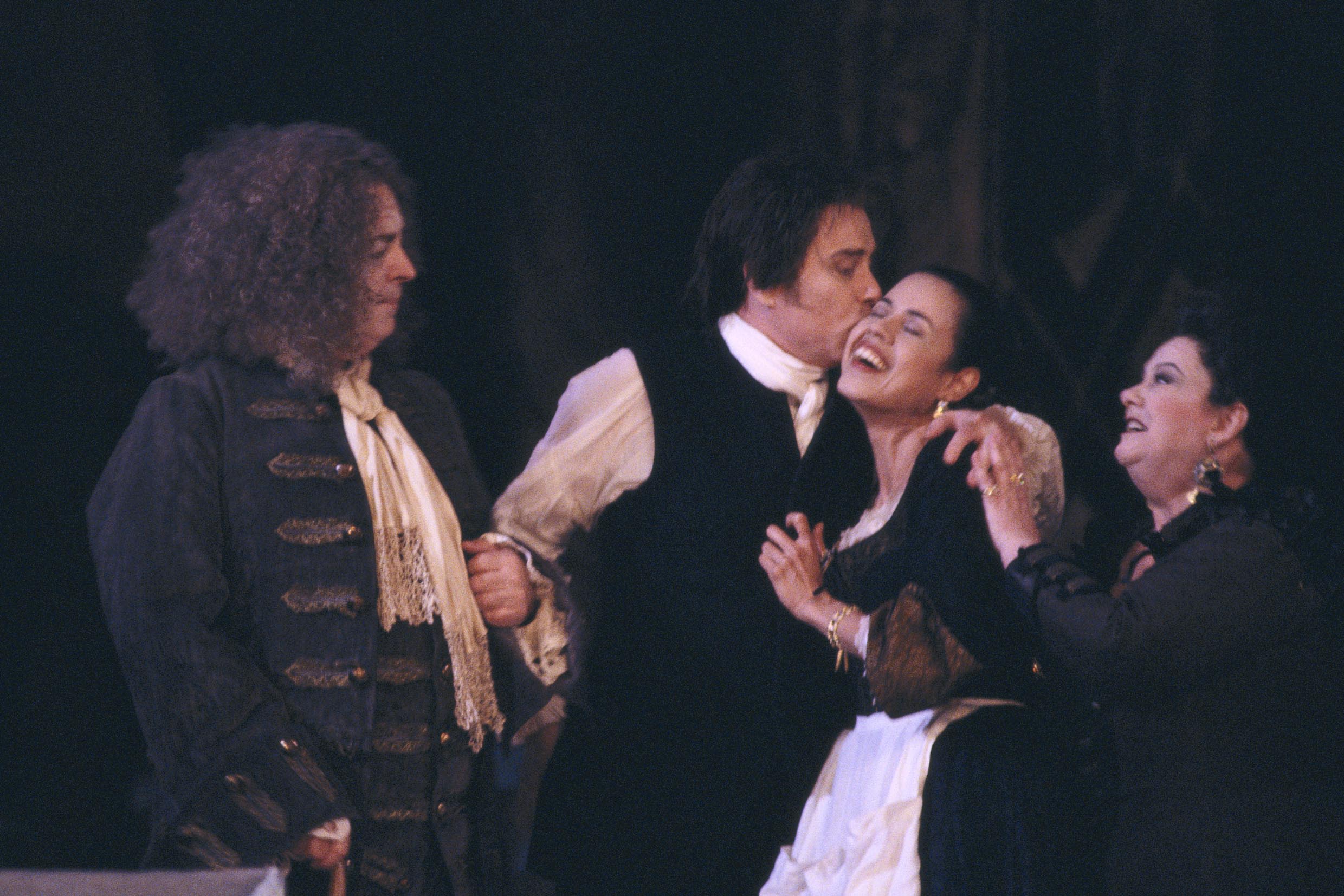A man kisses a woman on the cheek as they embrace. An older man and woman stand either side of the embracing couple. They are in a performance of The Marriage of Figaro.