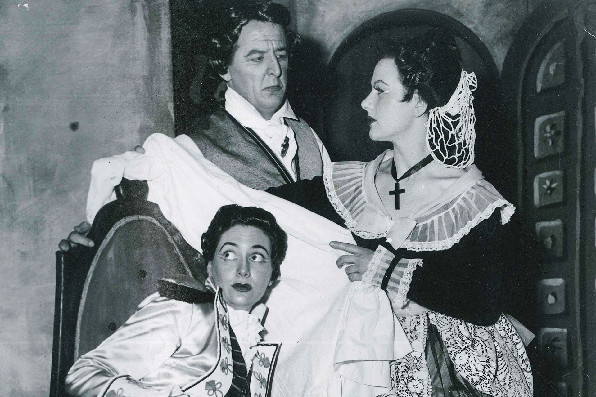A man and two women sit around a chair in period costume, in character for a performance of The Marriage of Figaro.