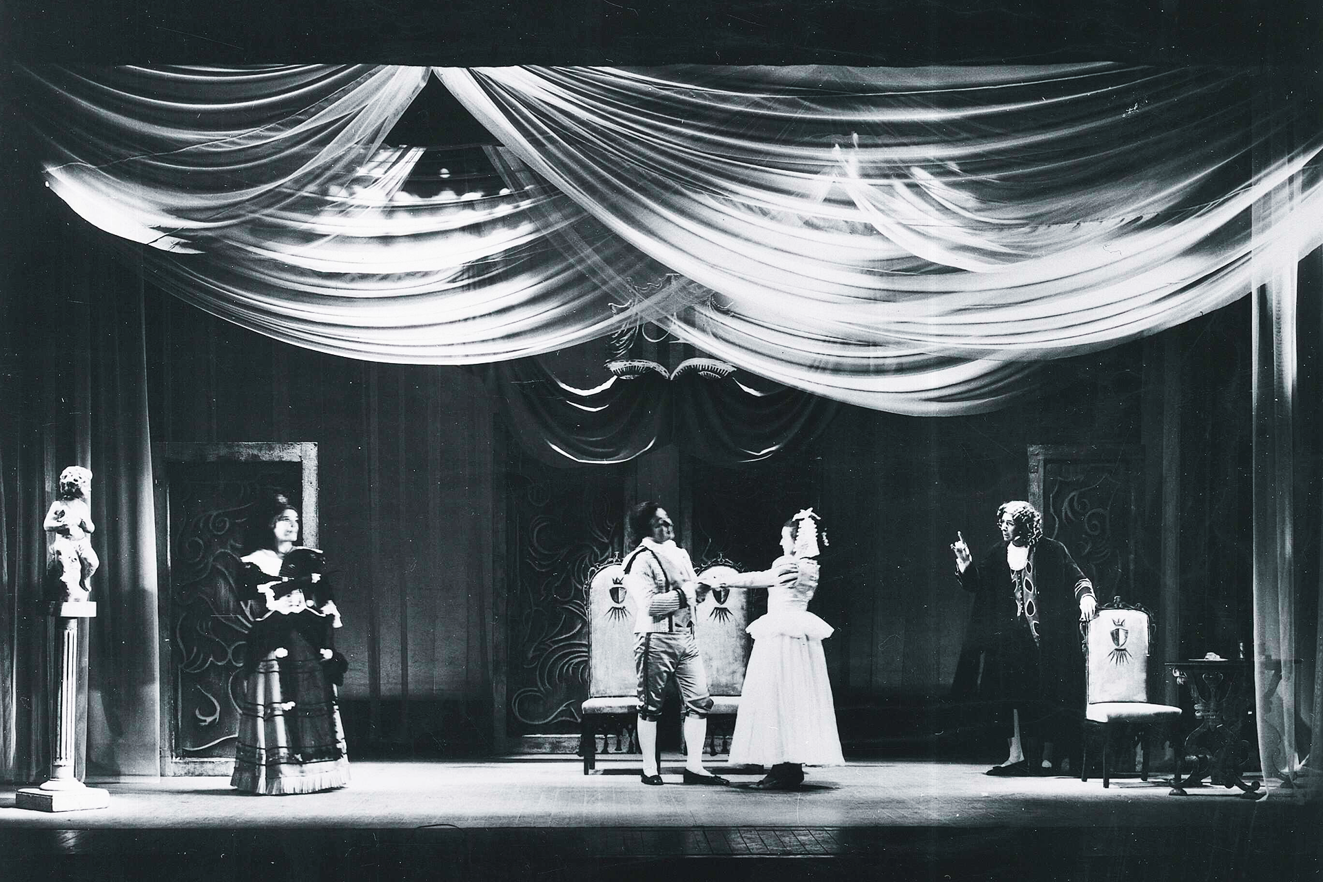A group of people in period costume appear on stage in a performance of The Marriage of Figaro. Sheer fabric is draped above the stage.