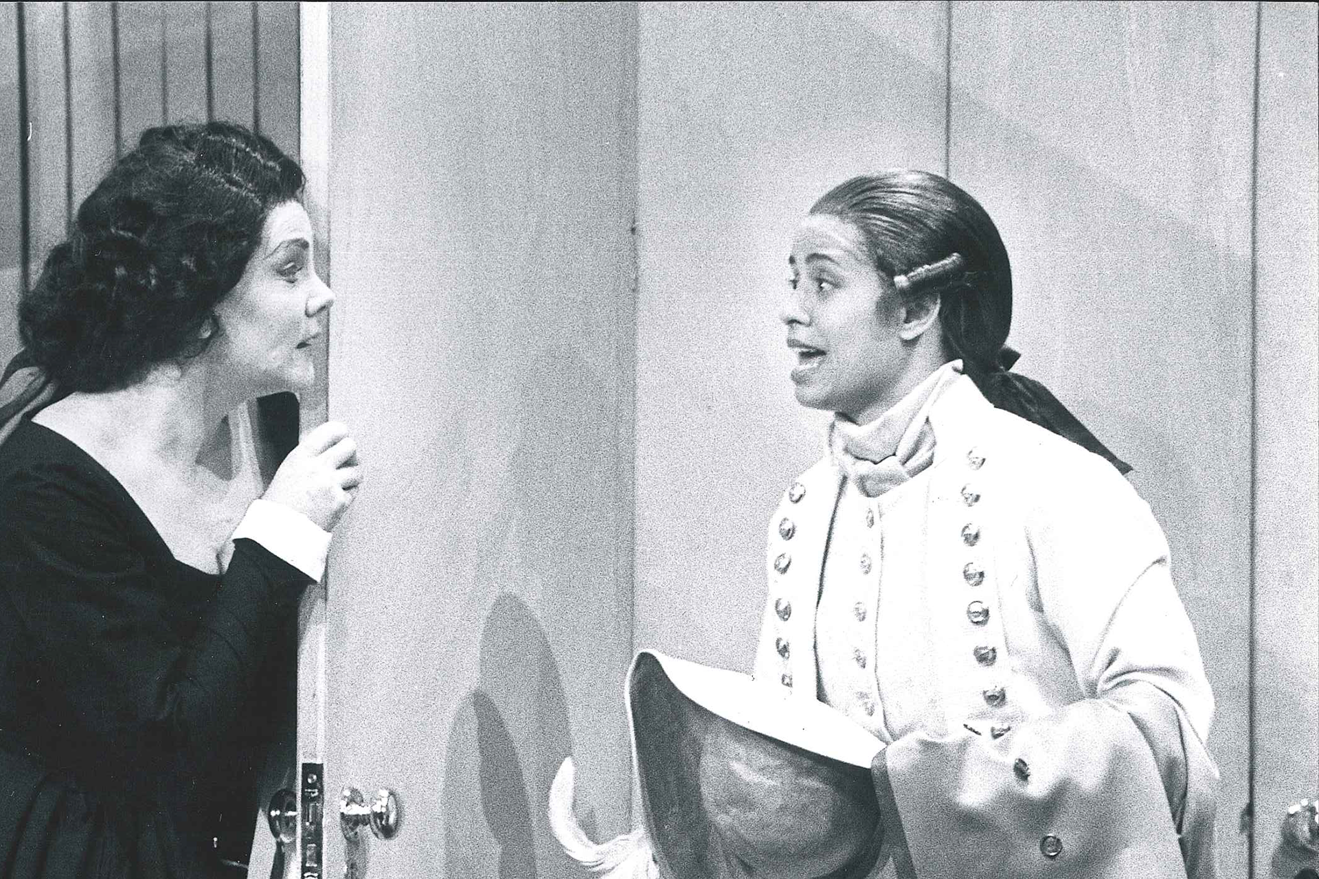 Two women stand either side of a door, one is dressed as a man. They are in a performance of The Marriage of Figaro.