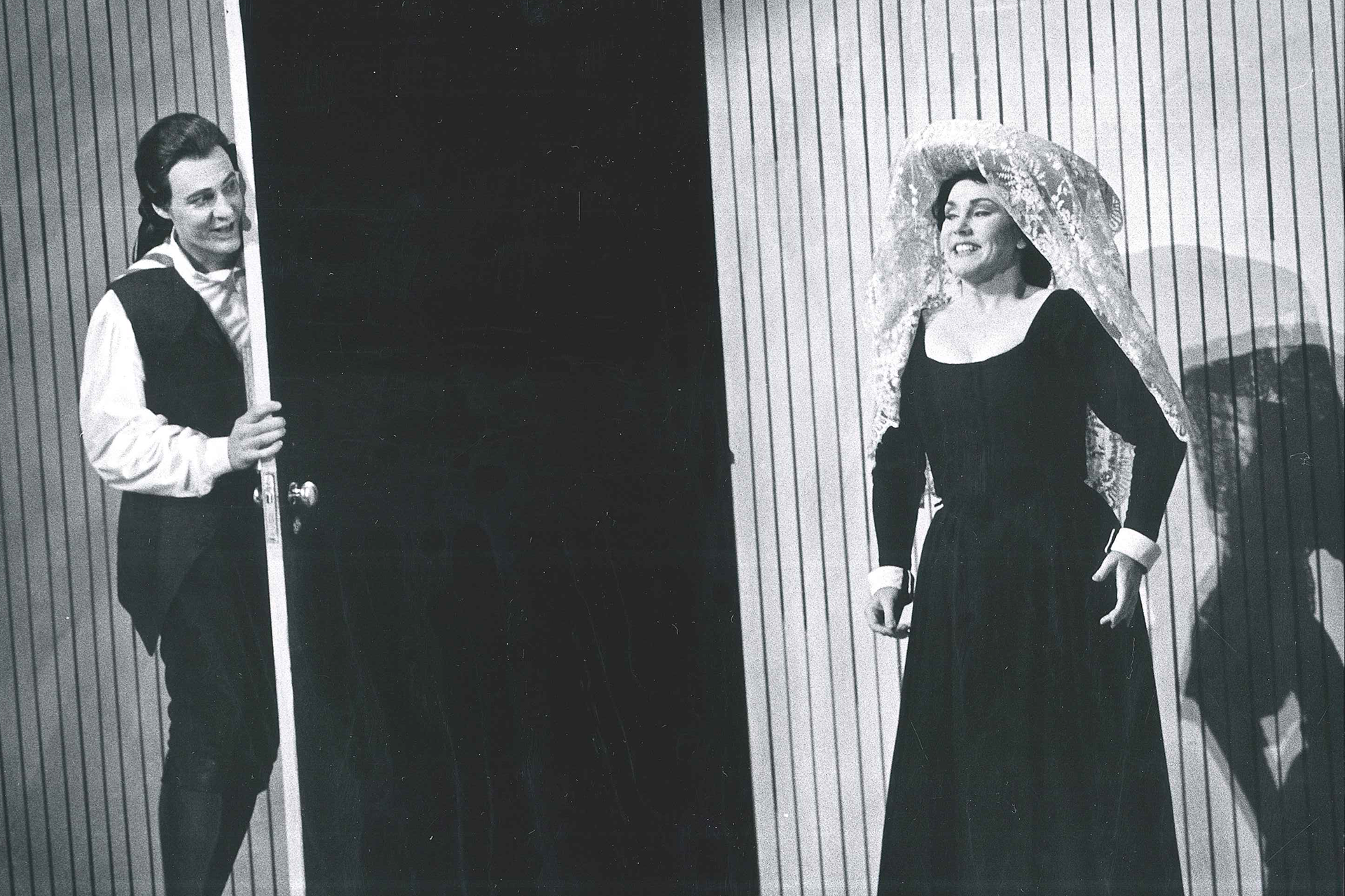 A man and a woman stand either side of a door, dressed in period costume, in a performance of The Marriage of Figaro.