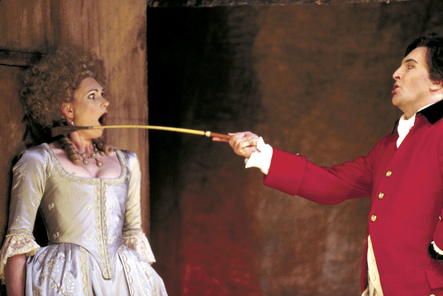 A man in a red jacket holds a riding crop to a woman's face in a performance of The Marriage of Figaro.