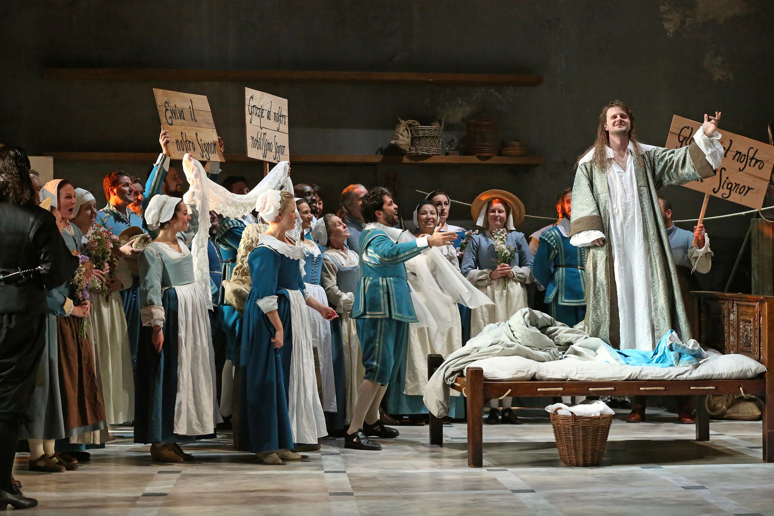A group of performers gather around a man standing on a bed in a performance of The Marriage of Figaro.