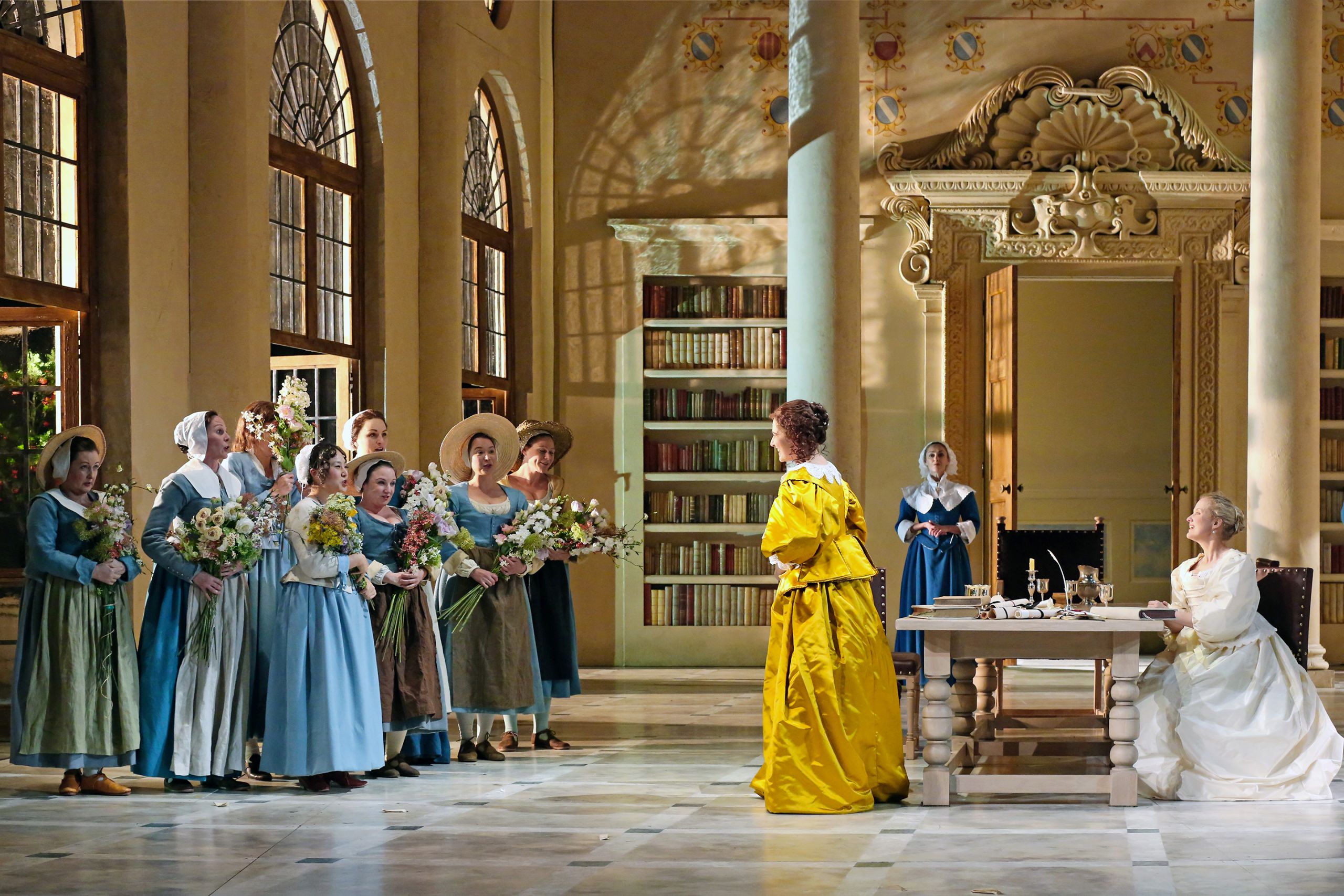 A group of singers in blue costumes bring flowers to a woman in a yellow gown in a performance of The Marriage of Figaro.