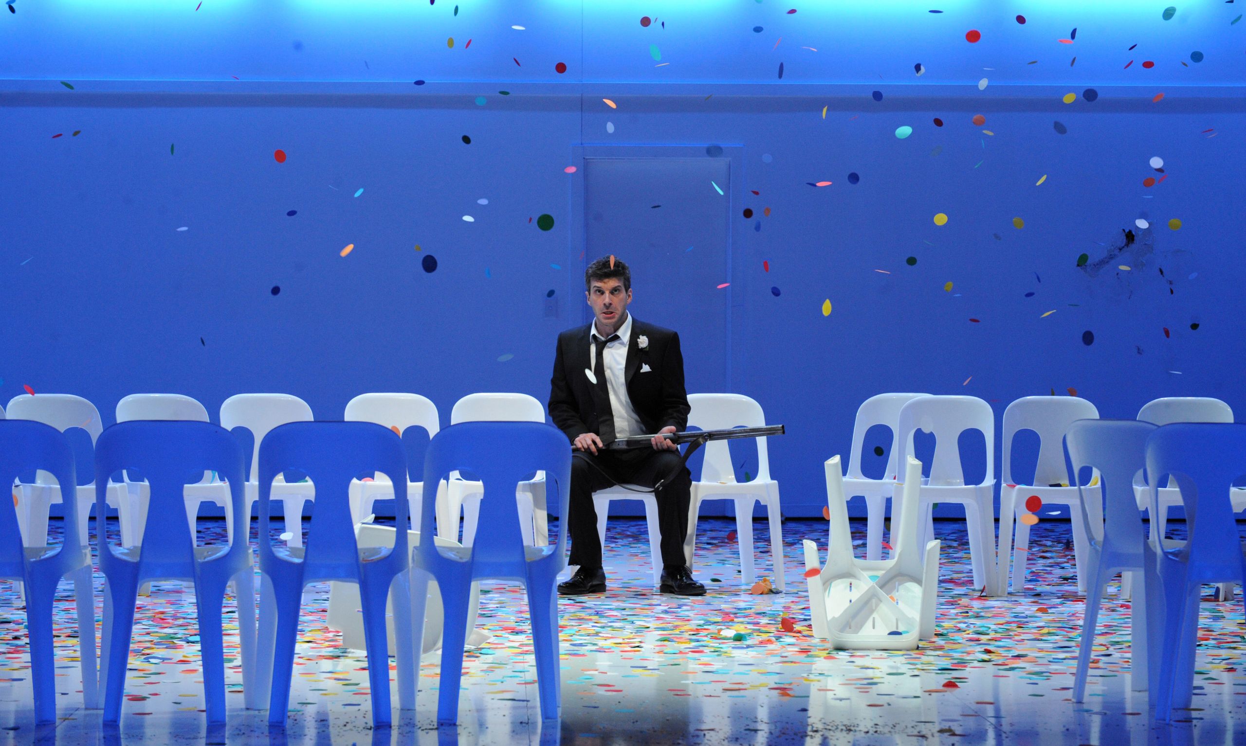 A man sits on a white plastic chair holding a rifle as colourful confetti falls around him during a performance of The Marriage of Figaro.