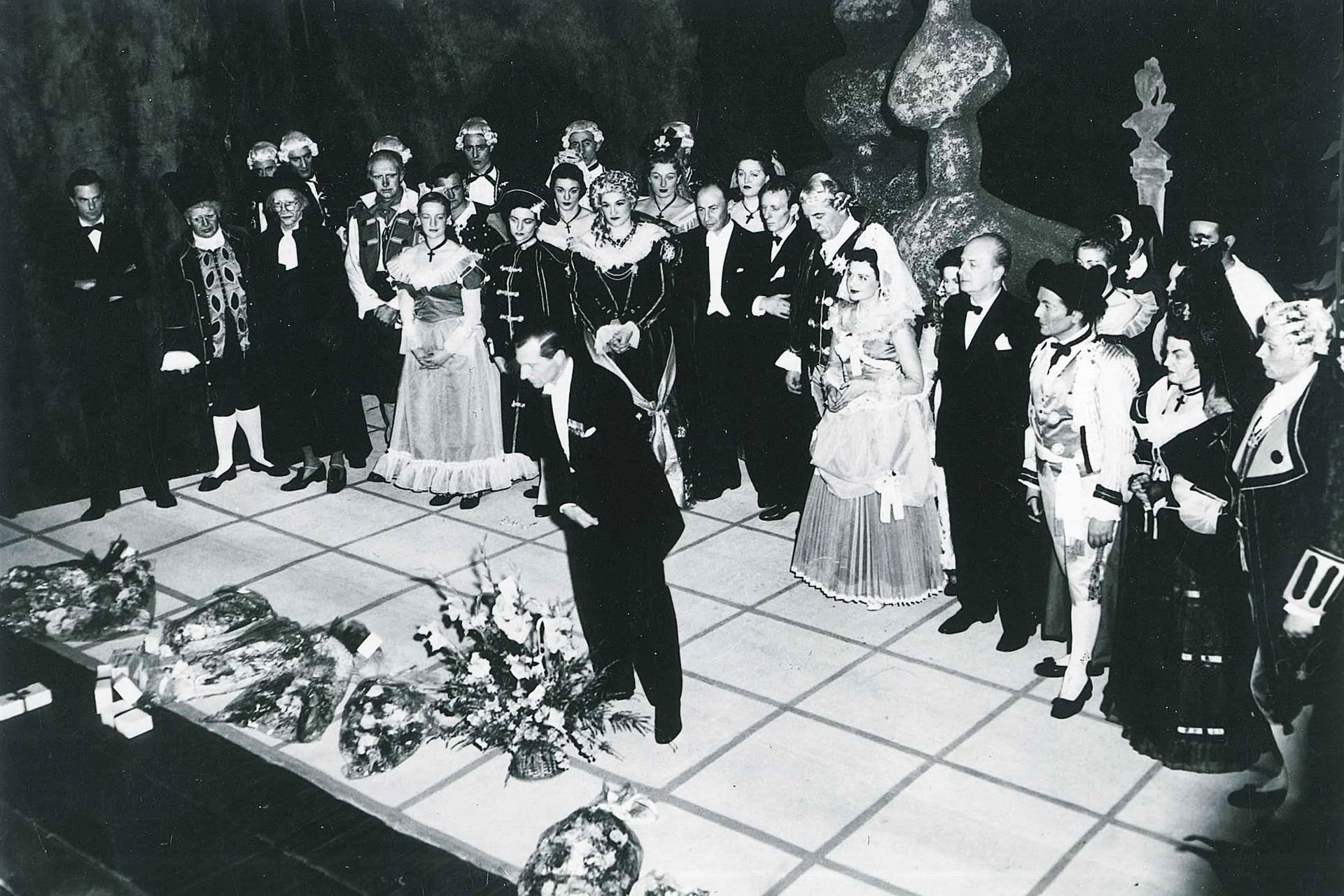 A man in a black tuxedo takes a bow on stage in front of a chorus of costumed opera singers.