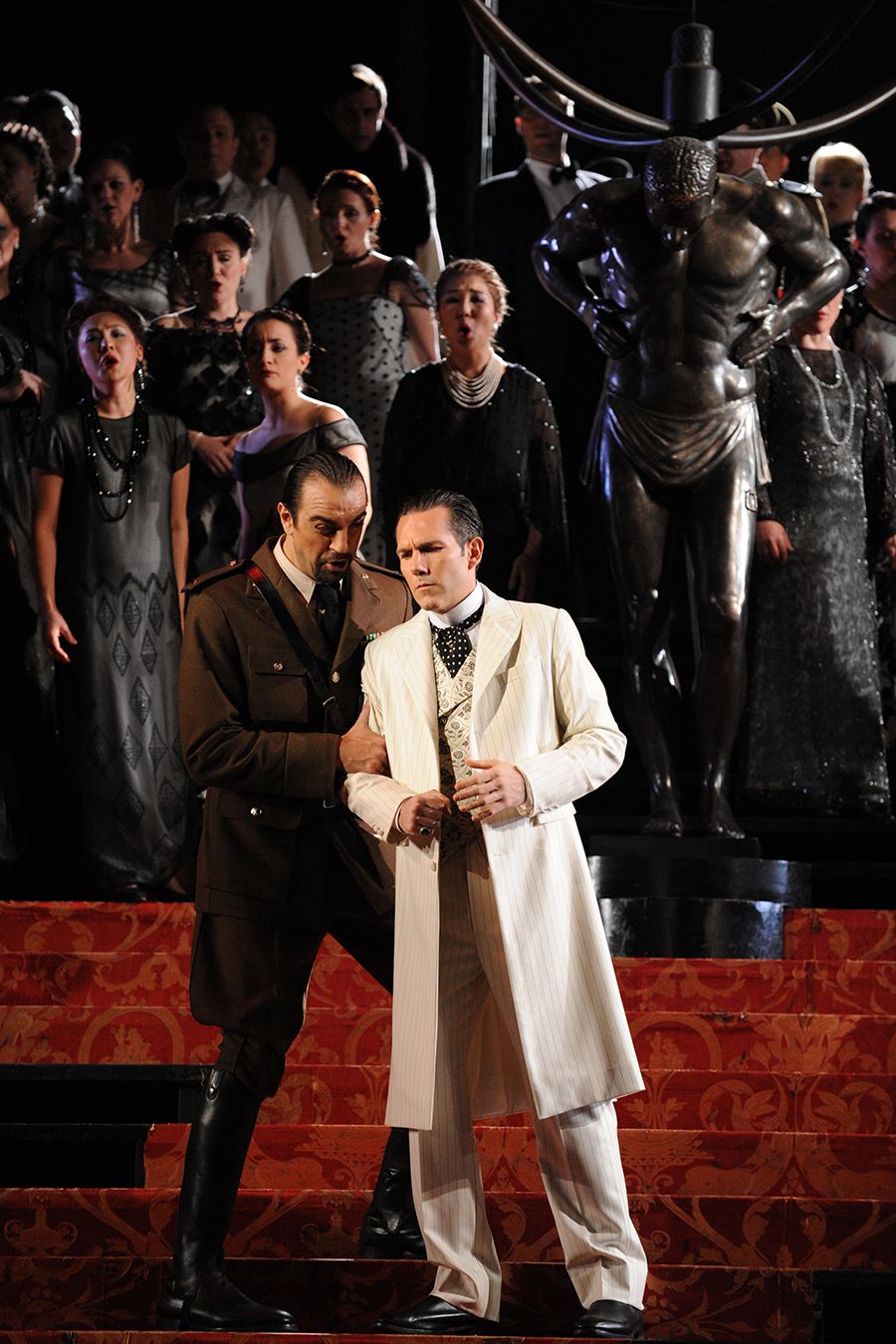 A man in a three-piece white suit stands with a man in military costuming on stage.