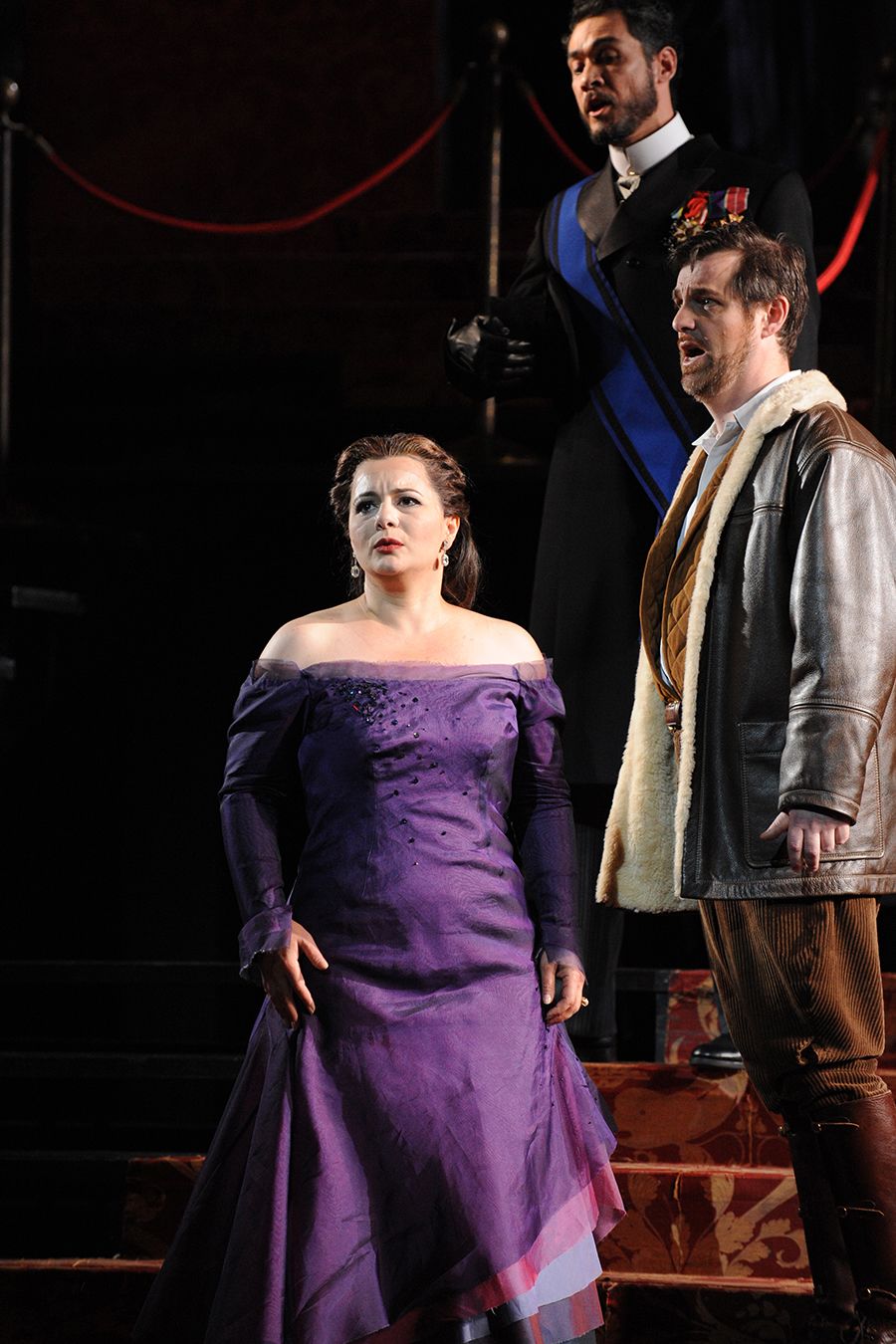 A woman stands in a purple gown with on red staircase with two men in formal wear behind.